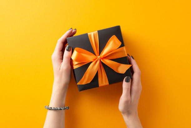 用万圣节礼物来取悦亲近的人。高角度看照片的女人的手拿着黑色的礼物盒与弓在橙色孤立的背景与copyspase广告或文字图片下载