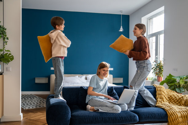 母亲在笔记本电脑上工作，而她活泼吵闹的孩子们在玩枕头，玩得很开心图片下载