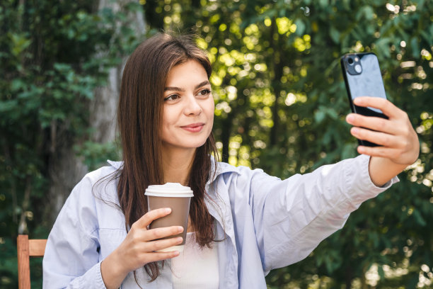 一位年轻女子拿着一杯咖啡，在森林的背景下自拍。图片下载