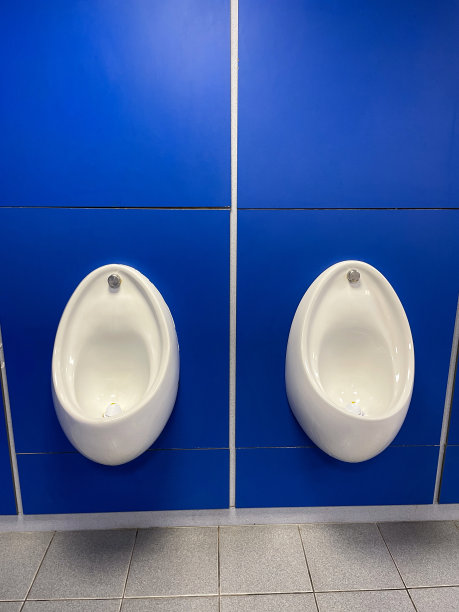 公共厕所的两个小便池图片，白色陶瓷小便池，蓝色背景飞溅，干净卫生的小便池，墙板内有排水管和排水系统，瓷砖地板图片下载