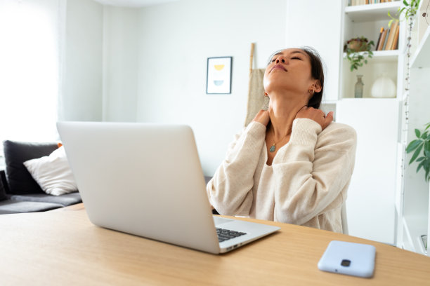 患有颈痛的亚洲女企业家。中国妇女在家用笔记本电脑按摩脖子。图片下载