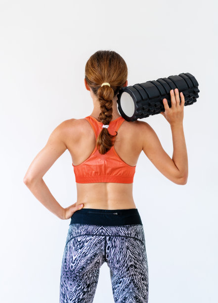 女健身教练站在工作室白色背景前的后视图照片，手里拿着泡沫滚筒。图片下载