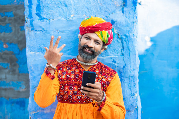 快乐的传统印度成熟男人穿着五颜六色的衣服，戴着头巾，使用智能手机，微笑的男人拿着安卓智能手机站在户外，蓝色的背景。节日。图片下载