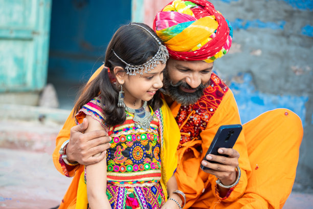 快乐的传统印度父亲和女儿穿着五颜六色的衣服，使用智能手机，微笑的胡子男人带着小女孩看视频，社交媒体互联网，安卓智能手机。图片下载