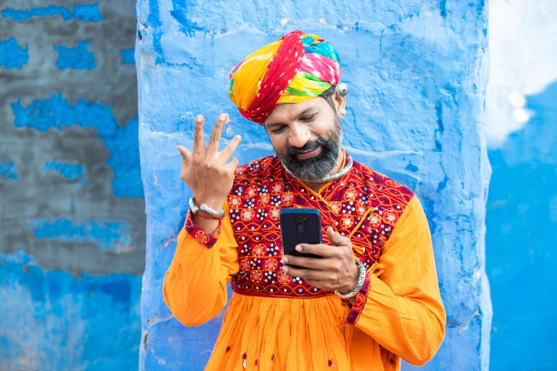 快乐的传统印度成熟男人穿着五颜六色的衣服，戴着头巾，使用智能手机，微笑的男人拿着安卓智能手机站在户外，蓝色的背景。节日。图片下载