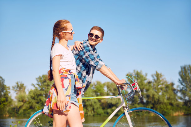 夫妇俩在夏日阳光下骑自行车散步的侧面照片，感到珍惜爱情。两个朋友看着彼此的爱走在河边的自行车。图片下载