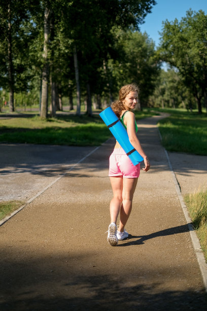 在公园里，一个年轻的卷发女人从后面拿着瑜伽垫和健身服的照片。图片下载