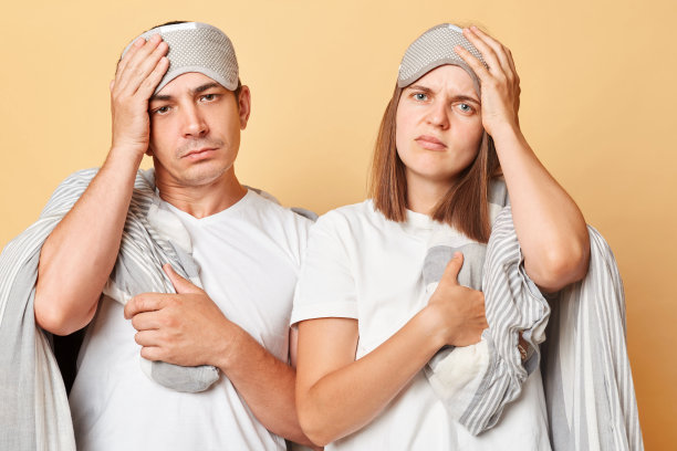 睡过头的一对夫妇戴着眼罩，用毯子裹着，隔绝在米色的背景下，早起头痛，需要休息。图片下载