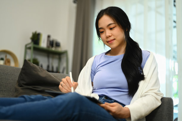 随意的亚洲女人坐在沙发上上网或在数码平板电脑上画画。图片下载