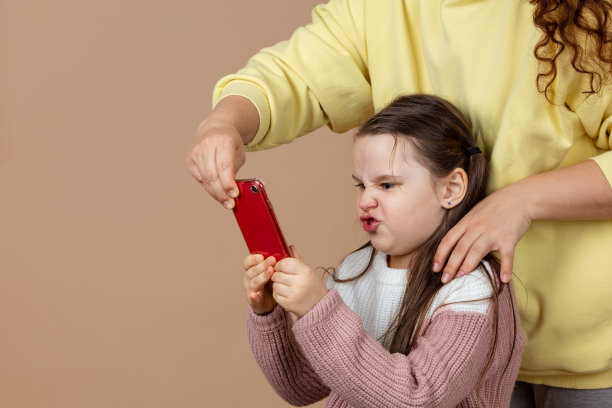 父母从女孩手中拿手机的照片，米色背景。不满的女儿拿着电话反击。预防儿童电话成瘾的概念，无手机恐惧症图片下载
