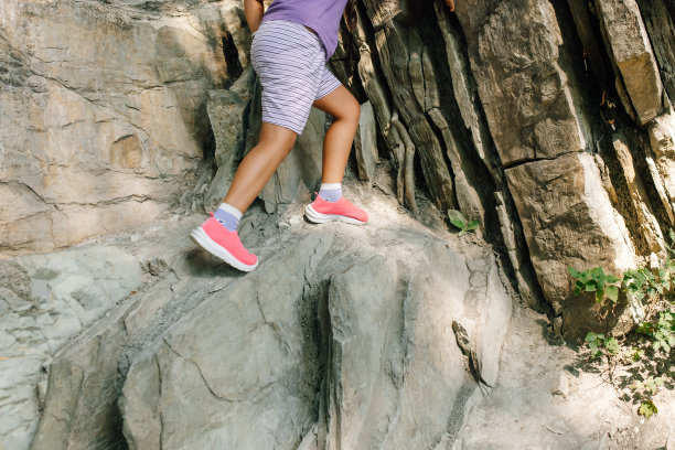 孩子的腿在山上爬上岩石的照片，侧面。穿着运动短裤和运动鞋的小女孩沿着危险的路线走下陡坡。旅游、滑坡、爬山、远足、运动图片下载
