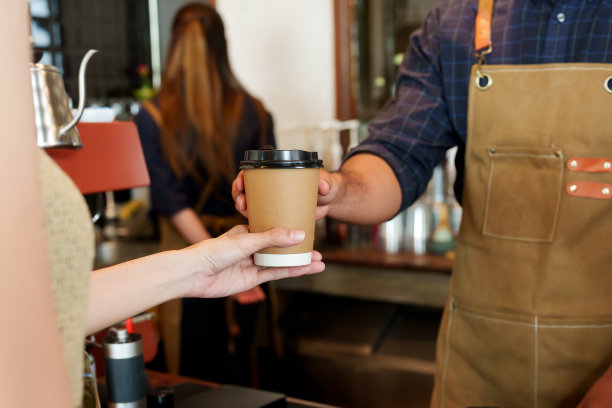 棕色的咖啡杯，黑色的咖啡杯盖正在被送到顾客手中。顾客从咖啡店老板那里收到咖啡在后台，他正在处理从其他顾客那里收到的订单图片下载