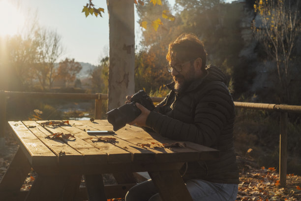 秋天，一名男子在户外用单反大相机坐在木桌上。公园里的黄叶。摄影师检查照片反射。户外休闲活动的人。现代的工作图片下载