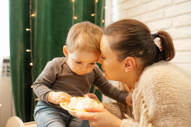 可爱的年轻女子亲吻幼童的家庭照片。母亲正在给儿子喂奶，拿着盘子靠近窗帘。图片下载