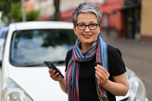 一个成熟的白人女性，一个快乐的司机，租了一辆有保险的新车，在户外用她的智能手机交流。图片下载