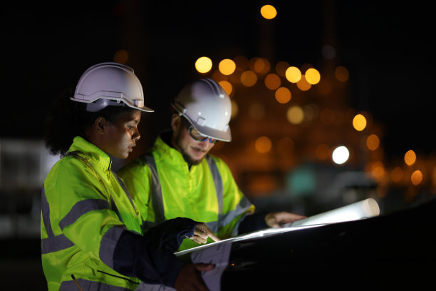 图为两名身穿绿色安全夹克的系统工程师在夜间在汽车引擎盖上工作时阅读图纸图片下载