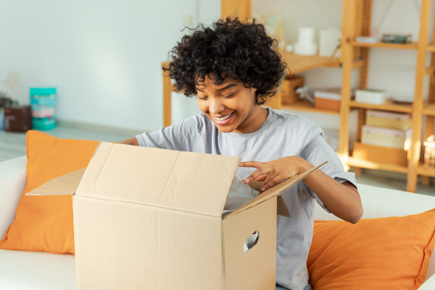 非洲女孩打开包裹，看着盒子。快乐的女人打开纸箱。女性拿着包裹，看着已经送达的物品。满意的客户积极反馈，快速快递交货。图片下载