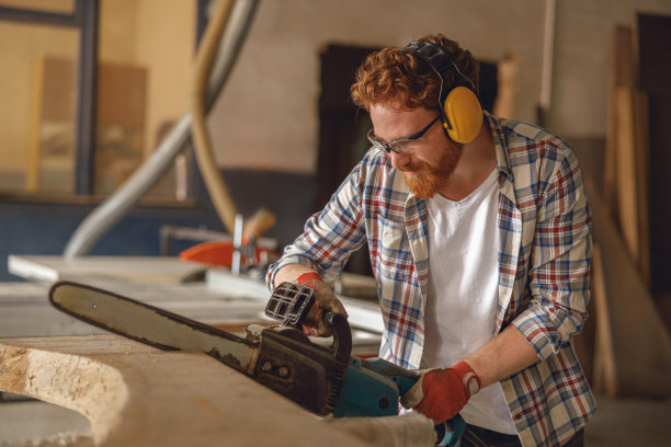 在木工车间，戴眼镜的木工正在用链锯锯木头。高质量照片图片下载