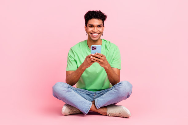 有趣的正面男子全身照片，穿着时髦的t恤裤子，坐在地板上，拿着智能手机，孤立在粉红色的背景上图片下载