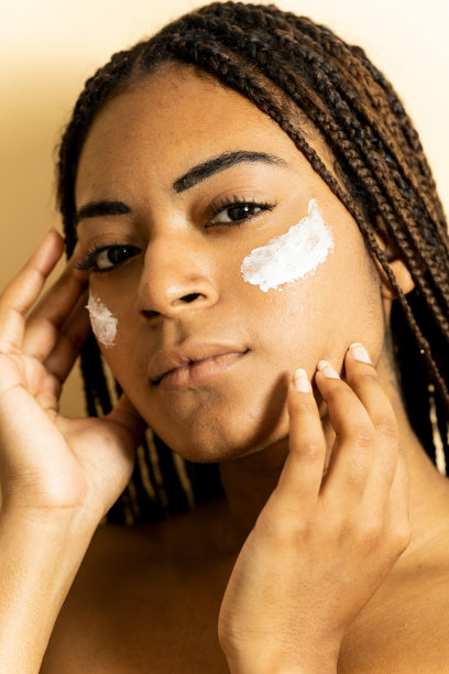 一个扎着辫子的非洲女孩的垂直特写照片，在棕色的摄影棚背景下摆姿势，她脸上涂着奶油，严肃地看着相机。防晒概念。图片下载