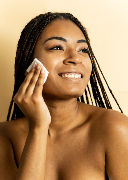 一个深色皮肤的女孩的垂直照片，她梳着辫子，在棕色的工作室背景下摆姿势。这名非洲妇女一边用一块布卸妆，一边看向一边。卸妆的概念。图片下载