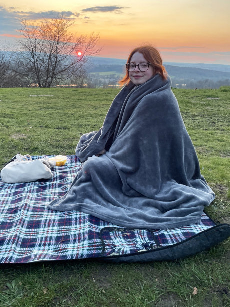 特写照片中，迷人的红发女子裹着毛茸茸的灰色毯子，坐在为淡季野餐准备的野餐毯上，日落时分在公园草地上浪漫约会，重点放在前景上图片下载