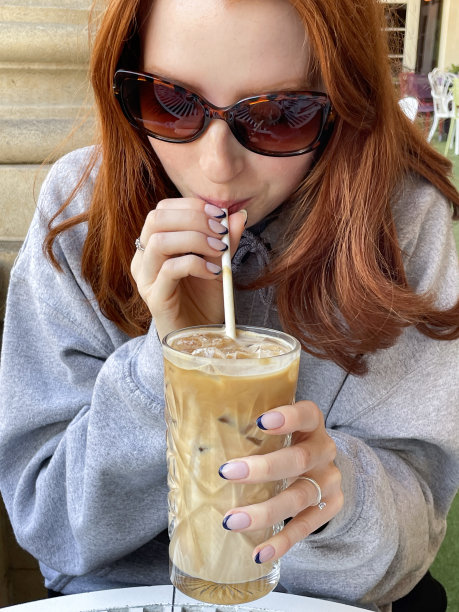 特写照片中，迷人的红发女子戴着墨镜，坐在铁制餐厅的露天餐桌前，手里拿着一杯冰咖啡饮料，用纸质吸管啜饮，聚焦于前景图片下载