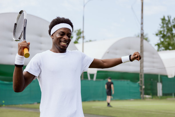 一个非洲人在打网球。他成功地战胜了对手，他满意地举起双手，微笑着。他拿着球拍，穿着白色t恤，头带和腕带图片下载