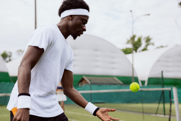 年轻的网球运动员在网球场上打网球。他抛出黄色的网球，准备击球。这名男子穿着白色t恤，带着绷带和腕带。他专注于比赛图片下载