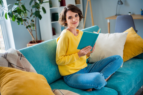 照片积极开朗的女士坐在沙发上阅读有趣的书为新智慧在现代客厅公寓图片下载