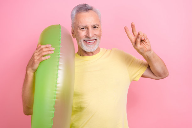 照片中，一个快乐有趣的退休老人在国外度假时，用粉红色背景的游泳救生圈展示了他的“v”手势图片下载