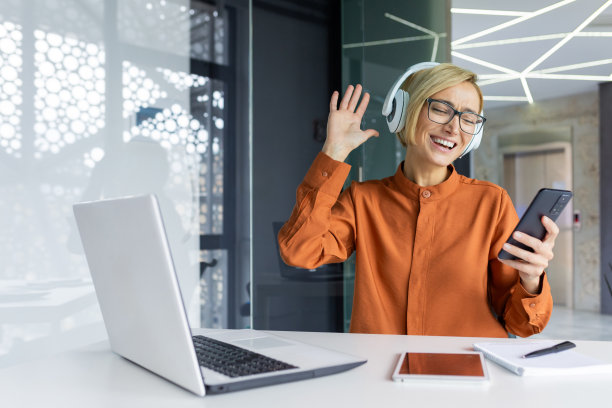 职场女性戴着耳机听音乐，在手机上使用应用程序，女员工欢快地跟着唱歌跳舞，拿着智能手机，坐在桌子上用笔记本电脑图片下载