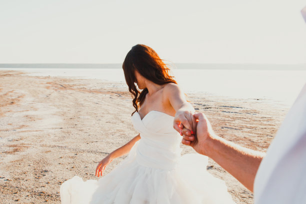 一对新人在海边举行婚礼。阳光明媚的夏日照片。新娘披散着头发，穿着露肩礼服，握着新郎的手。海洋的浪漫。海边的爱情故事。沙子，水和地平线。蜜月。图片下载