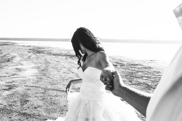 一对新人在海边举行婚礼。阳光明媚的夏日照片。新娘披散着头发，穿着露肩礼服，握着新郎的手。海洋的浪漫。海边的爱情故事。沙子，水和地平线。黑色和白色。图片下载