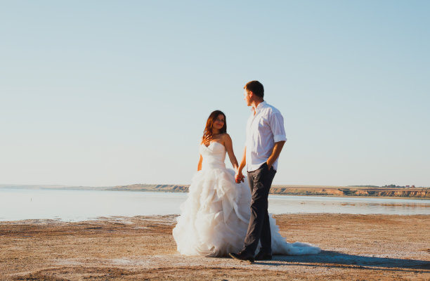 新婚夫妇在海边散步。阳光明媚的夏日照片。新娘披散着头发，穿着露肩裙。海洋浪漫的仪式。海边的爱情故事。沙子，水和地平线。蜜月。图片下载