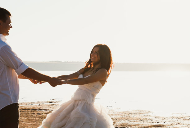 新婚夫妇在海滩上手拉手。阳光明媚的夏日照片。新娘披散着头发，穿着露肩裙。海洋浪漫的仪式。海边的爱情故事。沙子，水和地平线。蜜月。图片下载