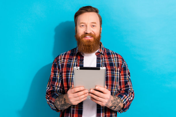 照片中，一个长着牙齿、笑容灿烂、留着时髦胡子、穿着格子衬衫的男人，拿着平板电脑，阅读电子邮件，孤立在蓝色背景下图片下载