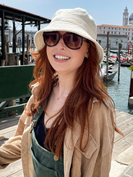 特写照片中，迷人的红发女子戴着墨镜和遮阳帽，站在威尼斯海滨的德格里河(Riva degli Schiavoni)上看着镜头，停泊在木桩上的船只，漂浮在水面上的贡多拉，意大利威尼斯，聚焦在前景上图片下载