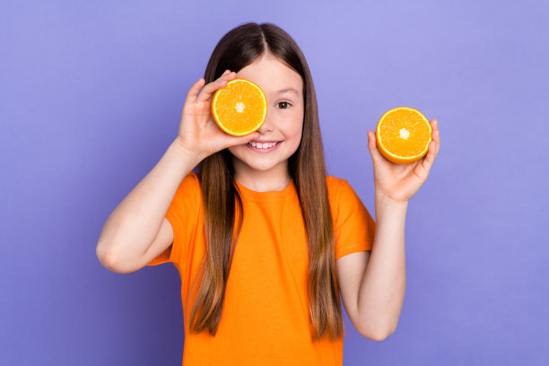 照片积极的学龄前女孩开朗的棕色头发穿橙色t恤遮住眼睛隐藏橙色水果孤立在紫色的背景图片下载