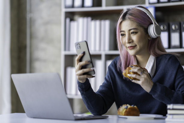 亚洲女商人坐着吃面包，看手机，读课间书，在办公室放松，愉快而美味地工作。图片下载