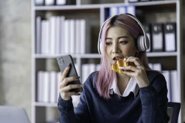 亚洲女商人坐着吃面包，看手机，读课间书，在办公室放松，愉快而美味地工作。图片下载