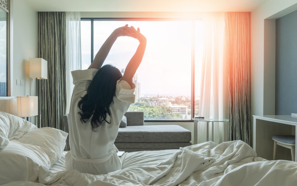 慵懒的一天，亚洲女性在周末早上从床上睡个好觉醒来，在舒适的卧室里休息，看着城市的景色，拥有快乐，工作与生活质量平衡的生活方式图片下载