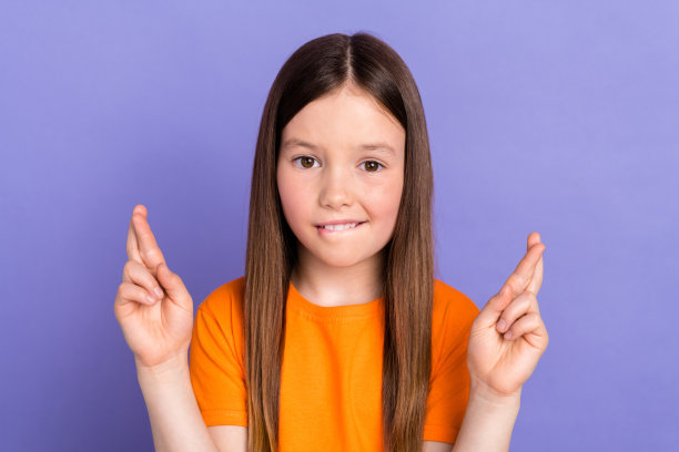 可爱的漂亮学生的照片，直发，穿着橙色t恤，手指折叠，在紫色的背景下许愿图片下载