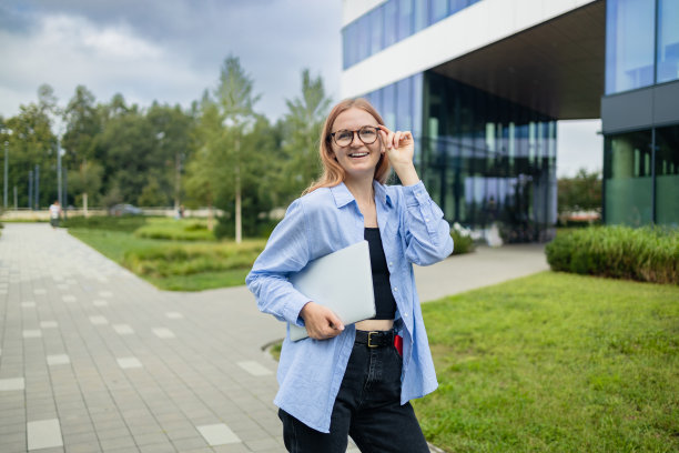 这位自信成功的年轻女性，穿着正式，拿着笔记本电脑站在户外，以商业中心为背景，微笑着看着镜头图片下载