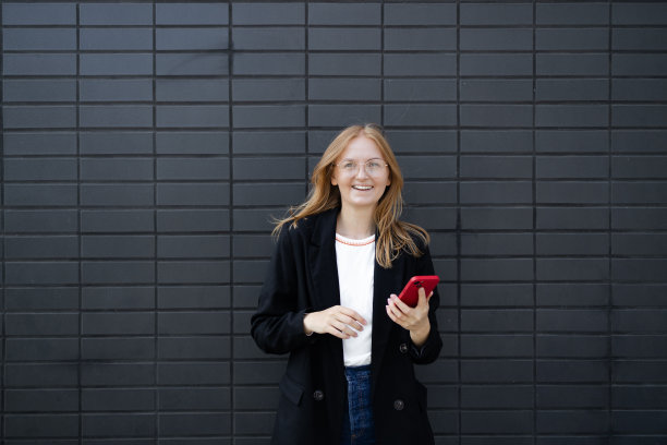 开朗的金发女商人独自站在灰色的墙壁背景上，手里拿着手机。微笑着可爱的年轻女子站在灰色背景上使用手机图片下载