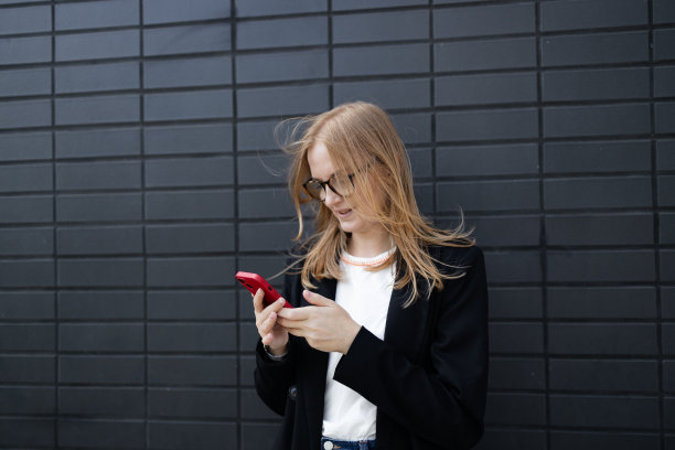 开朗的金发女商人独自站在灰色的墙壁背景上，手里拿着手机。微笑着可爱的年轻女子站在灰色背景上使用手机图片下载