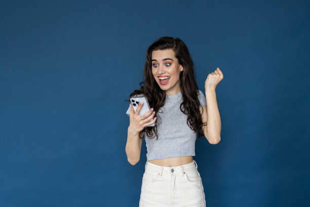 照片中，一个快乐而惊讶的年轻女子被隔离在蓝色的墙壁背景下，用手机做出了获胜的手势。图片下载