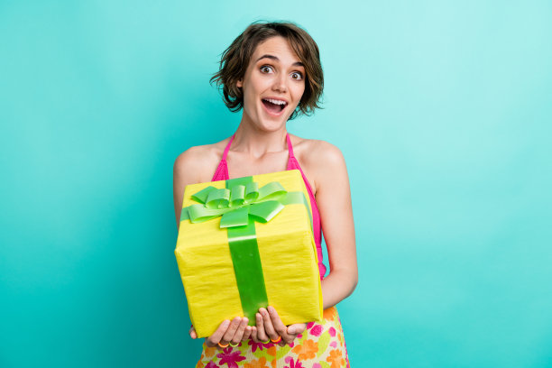 惊喜的照片，开朗的女孩张开嘴，穿着粉红色的时髦衣服，拿着大尺寸的生日盒子，孤立在青色背景上图片下载