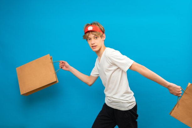 年轻的卖家穿着休闲的白色t恤，蓝色背景的工作室肖像上有一个纸袋。人们的生活理念。模拟复制空间。十几岁的孩子拿着装着买的东西的包图片下载