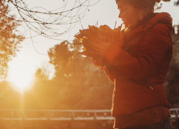 一位女士喜欢大自然，在秋天夕阳的金色时刻捧着枯叶。人与户外休闲活动。自由和宁静幸福的女性在公园。橙色图片配树图片下载